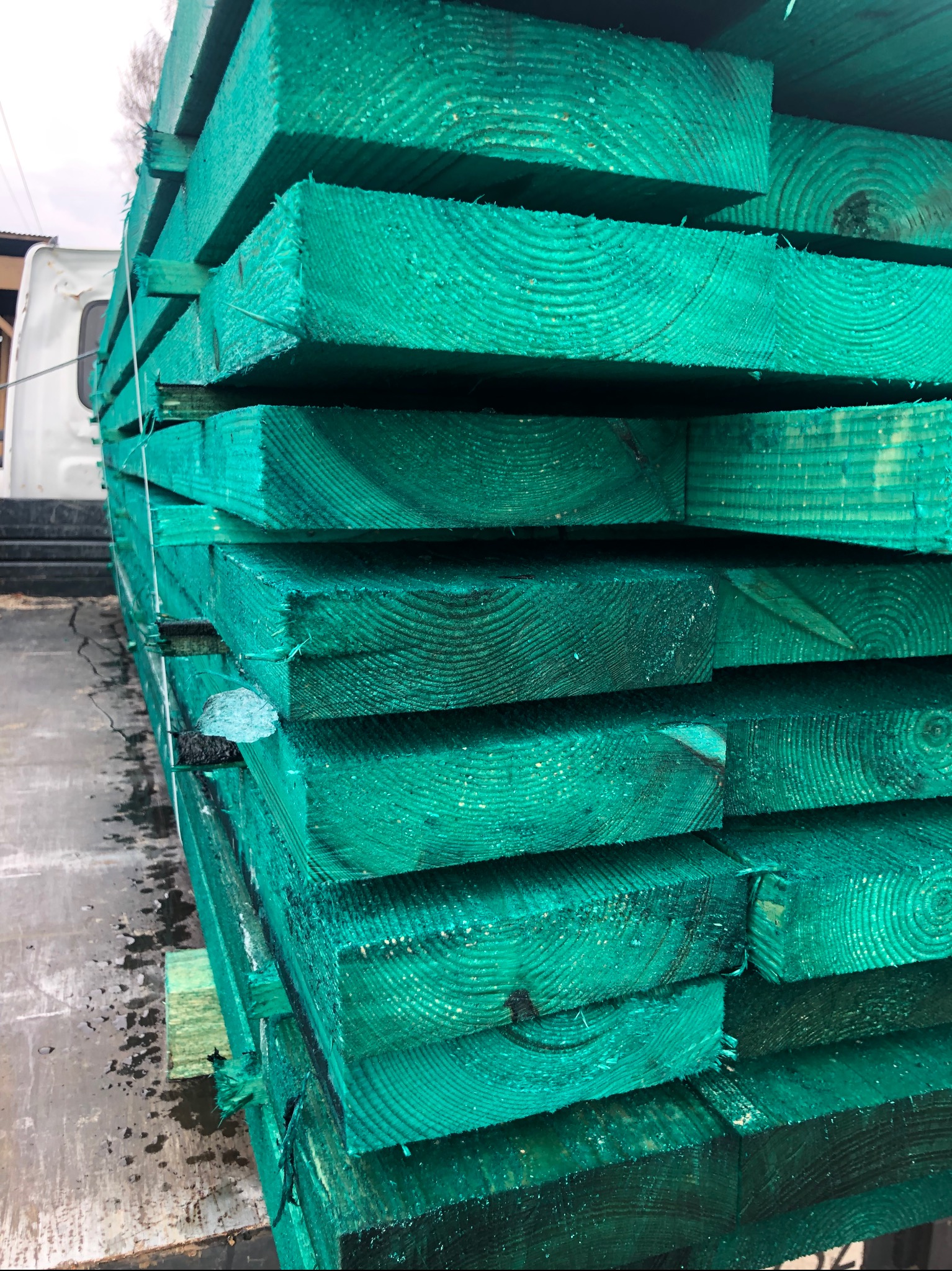 Stos impregnowanej na zielono tarcicy ułożonej na palecie, widoczny fragment białej ciężarówki w tle.