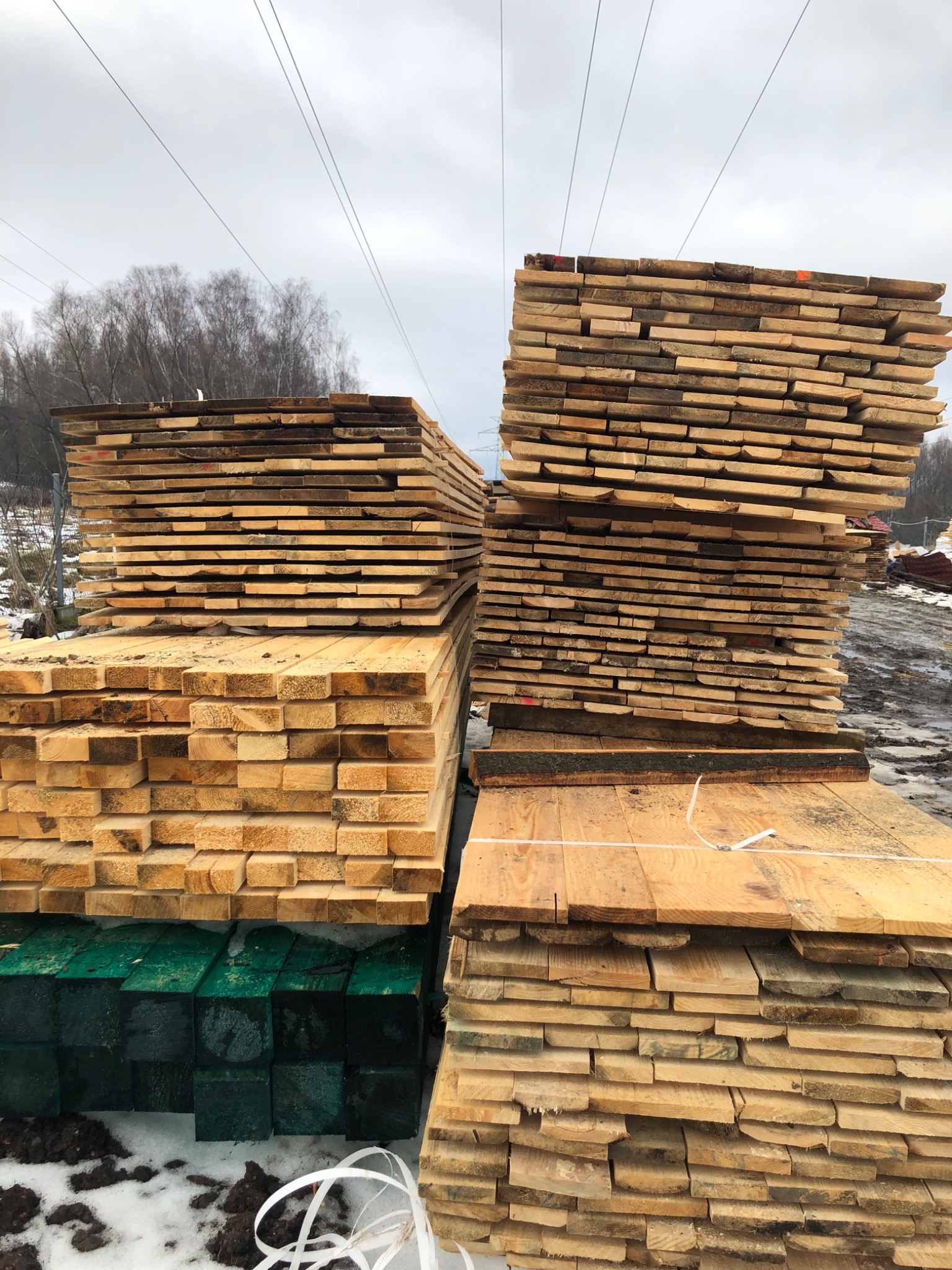 Stosy świeżo przetartej tarcicy budowlanej ułożone na zielonych, impregnowanych podkładach, widoczne ślady śniegu i błota na placu składowym, w tle drzewa i słupy wysokiego napięcia.