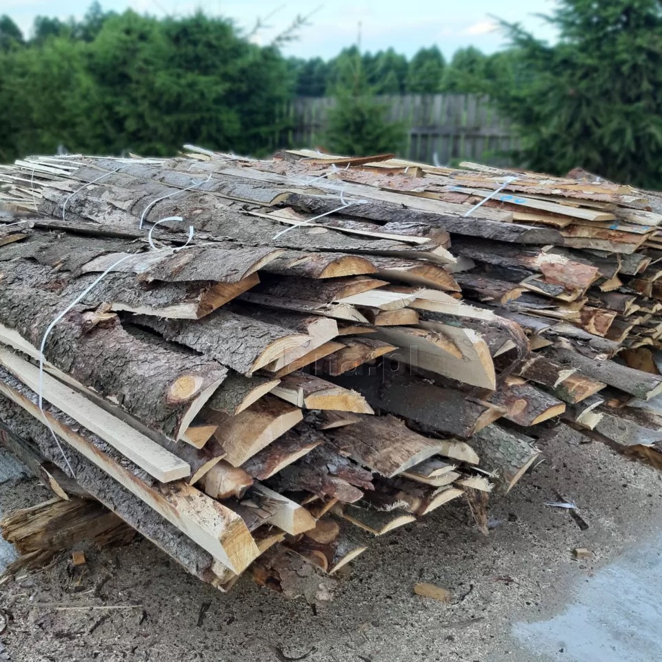 Stos tarcicy z korą, związany białym sznurkiem, na tle zielonych drzew i drewnianego płotu.