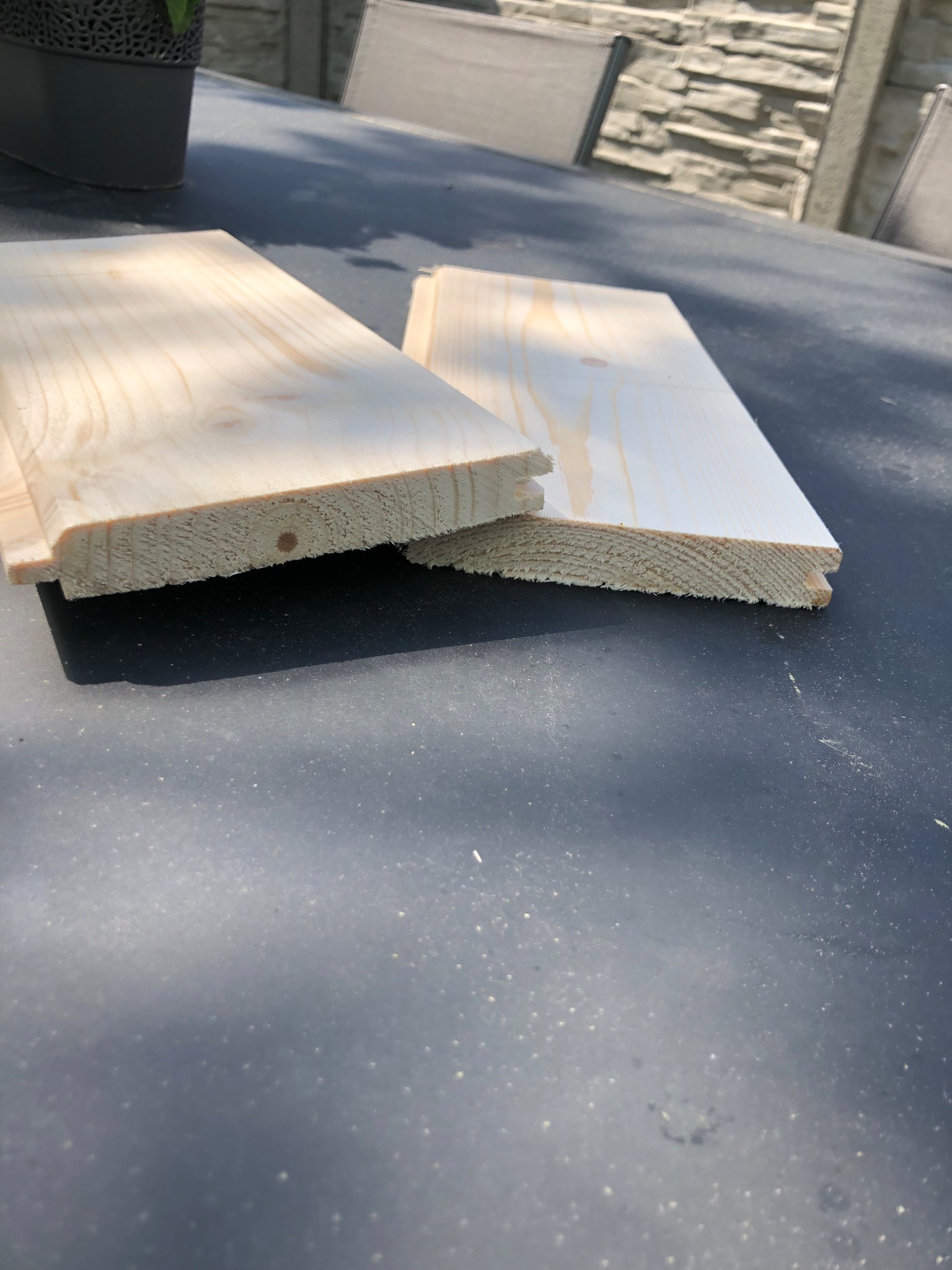Dwie deski z jasnego drewna, jedna z widocznym sękiem, leżące na szarym stole na zewnątrz, z fragmentem kamiennego ogrodzenia i krzesłami w tle.