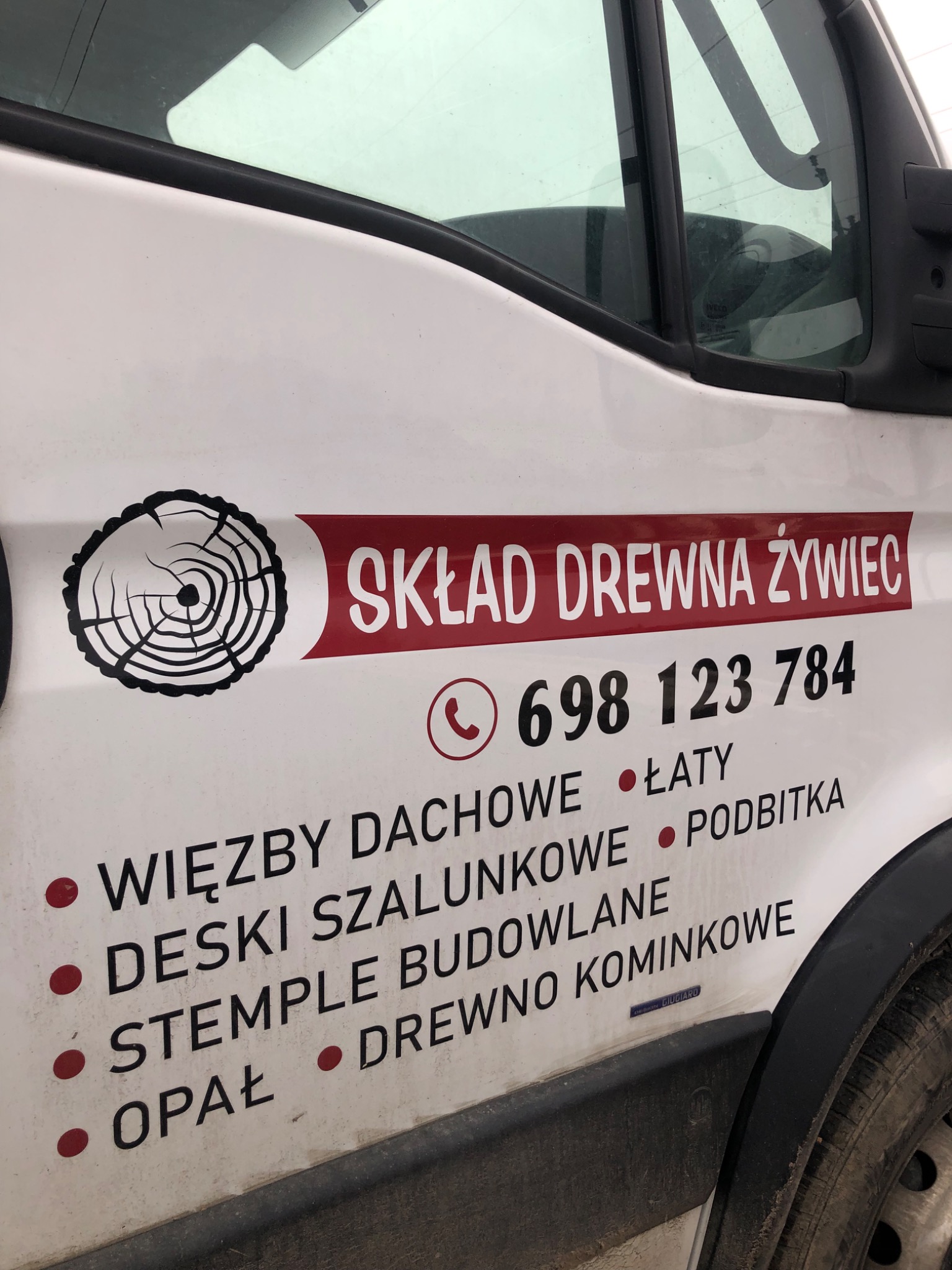 Bok białego samochodu dostawczego z reklamą składu drewna Żywiec, zawierającą logo z przekrojem pnia drzewa, numer telefonu i listę produktów: więźby dachowe, deski szalunkowe, stemple budowlane...