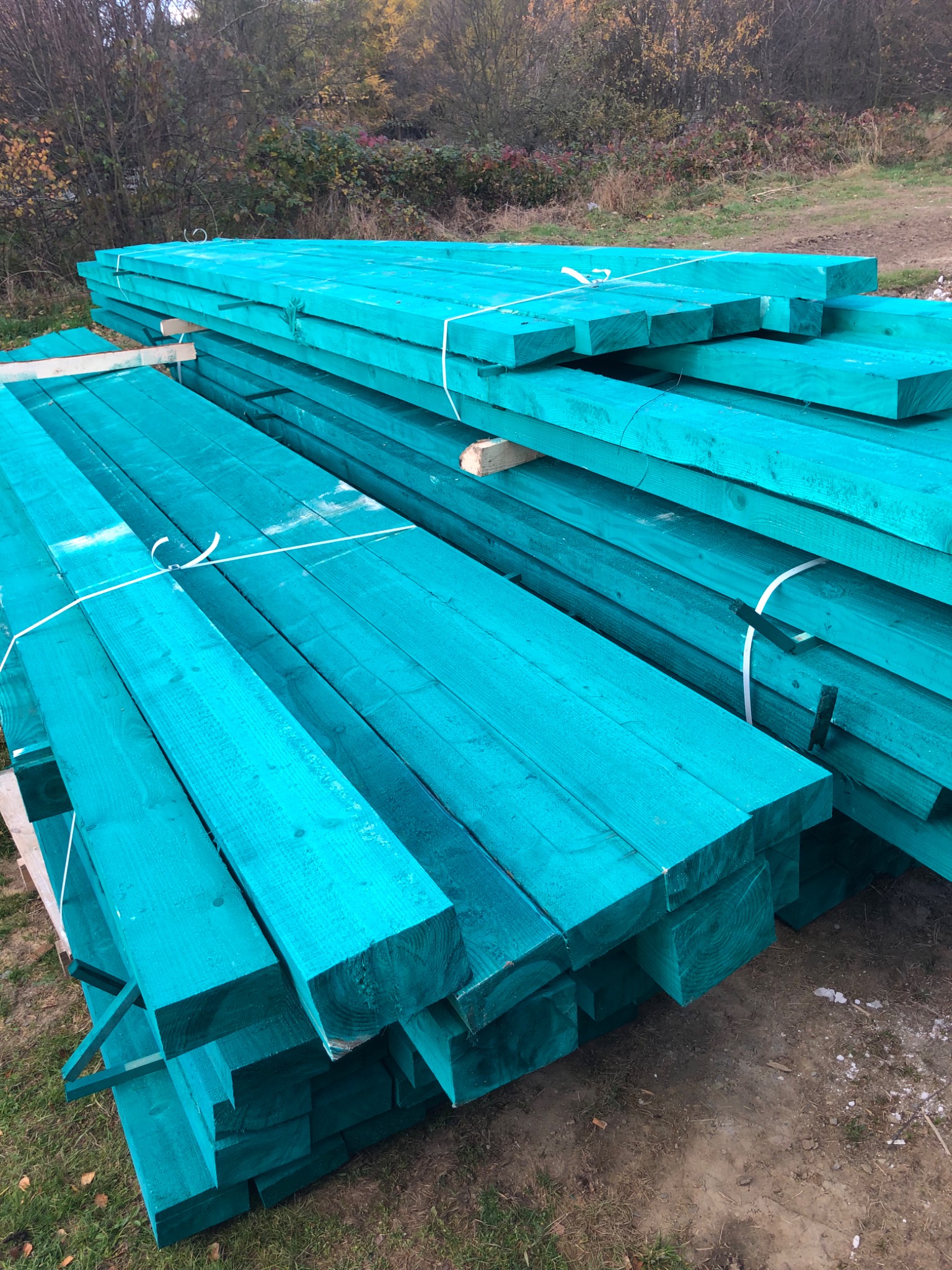 Stos turkusowej tarcicy budowlanej ułożonej na zewnątrz, zabezpieczonej białym sznurkiem, z widocznym tłem jesiennych drzew