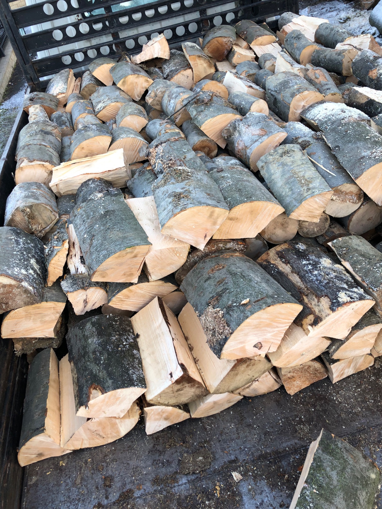 Stos pociętego drewna liściastego z korą, ułożonego w metalowej skrzyni transportowej, widoczne ślady śniegu.