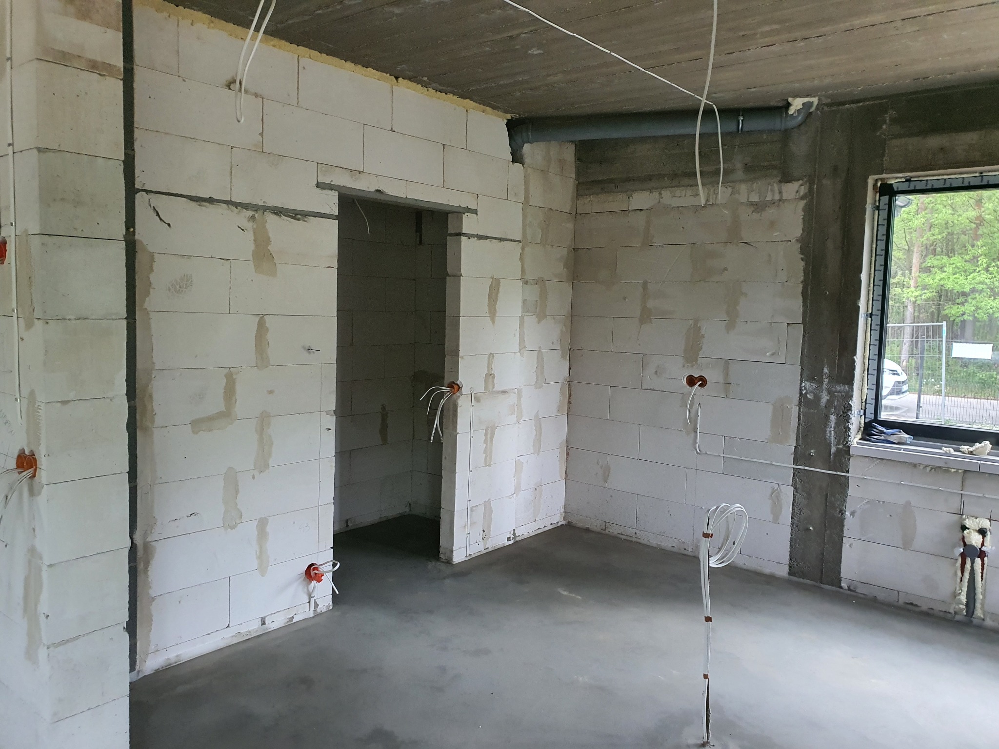 Wnętrze surowego budynku w trakcie prac instalacyjnych: widoczne ściany z betonu komórkowego, wystające przewody elektryczne w pomarańczowych puszkach, niezabudowane okno z widokiem na zieleń.