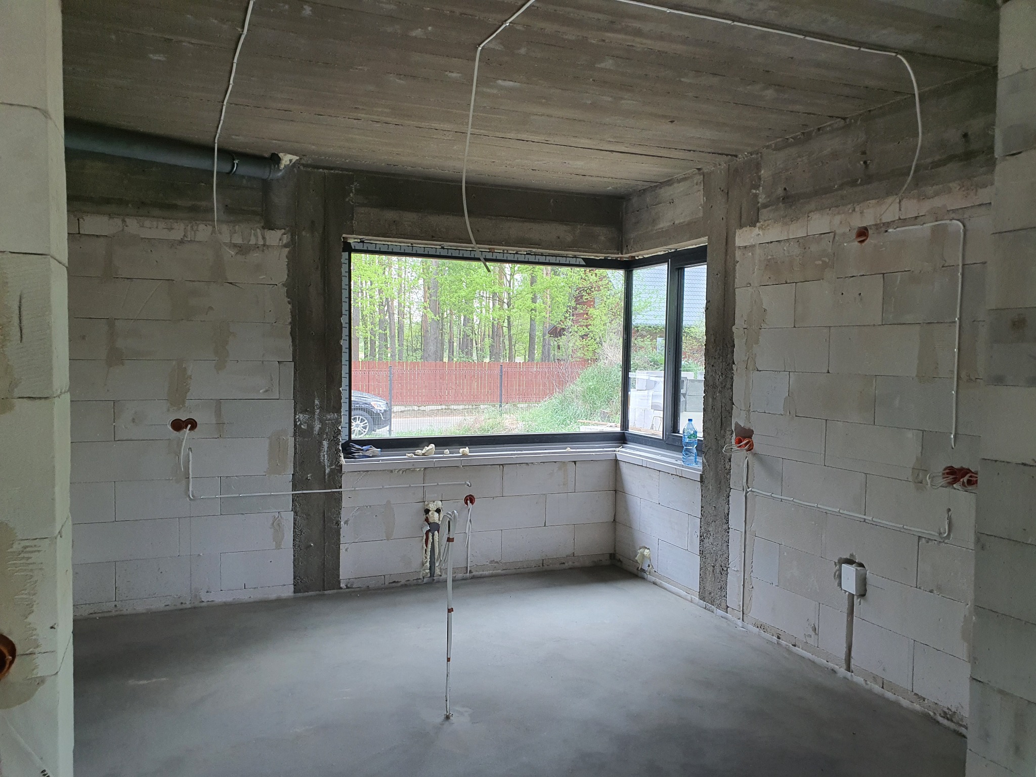 Surowy stan wnętrza z instalacją elektryczną: przewody na ścianach z betonu komórkowego, okno z widokiem na zieleń i woda mineralna na parapecie.