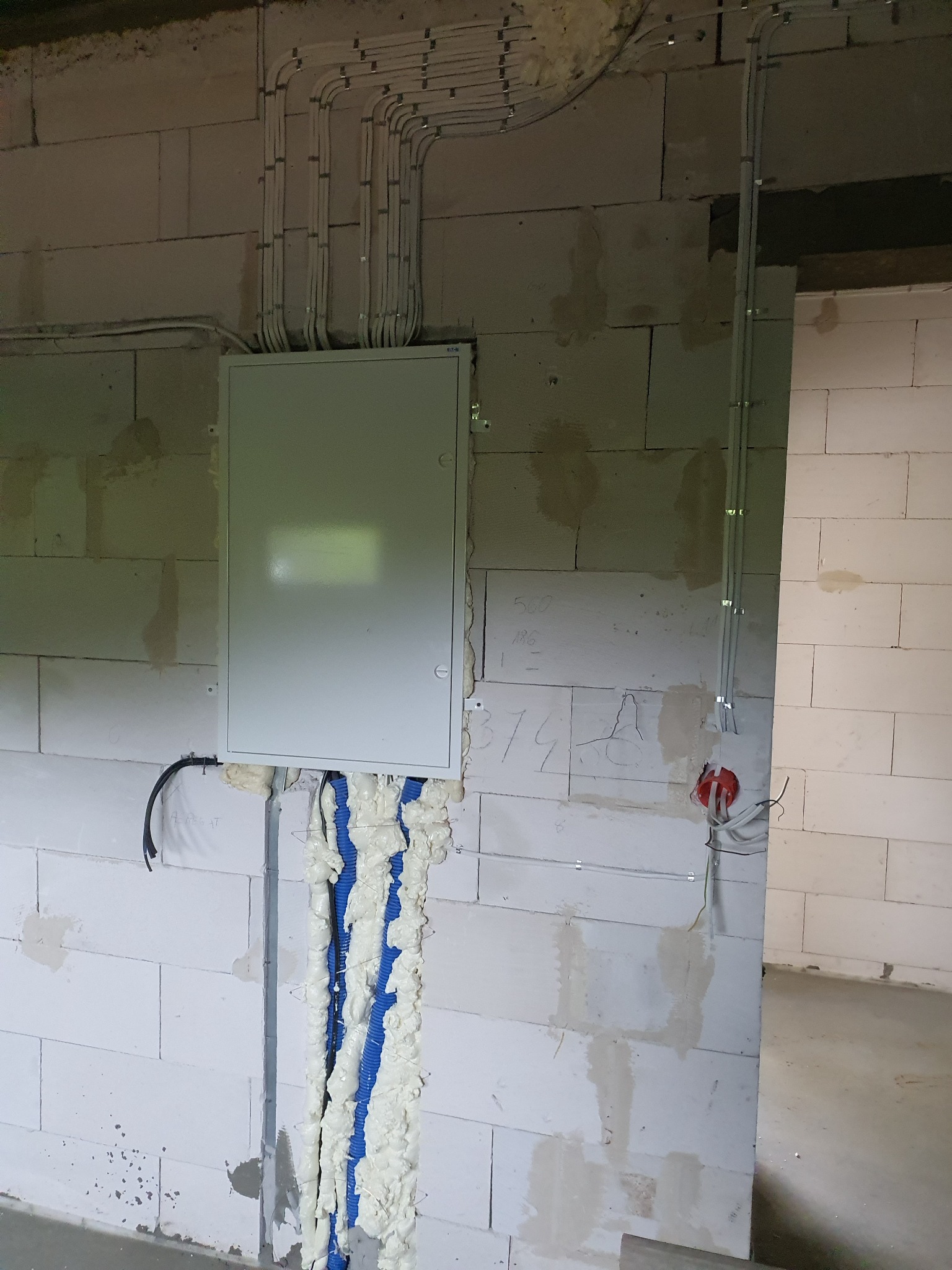Rozdzielnica elektryczna na surowej ścianie z betonu komórkowego, z widocznymi przewodami w rurkach PCV prowadzonych po ścianie i zabezpieczonych pianką montażową.