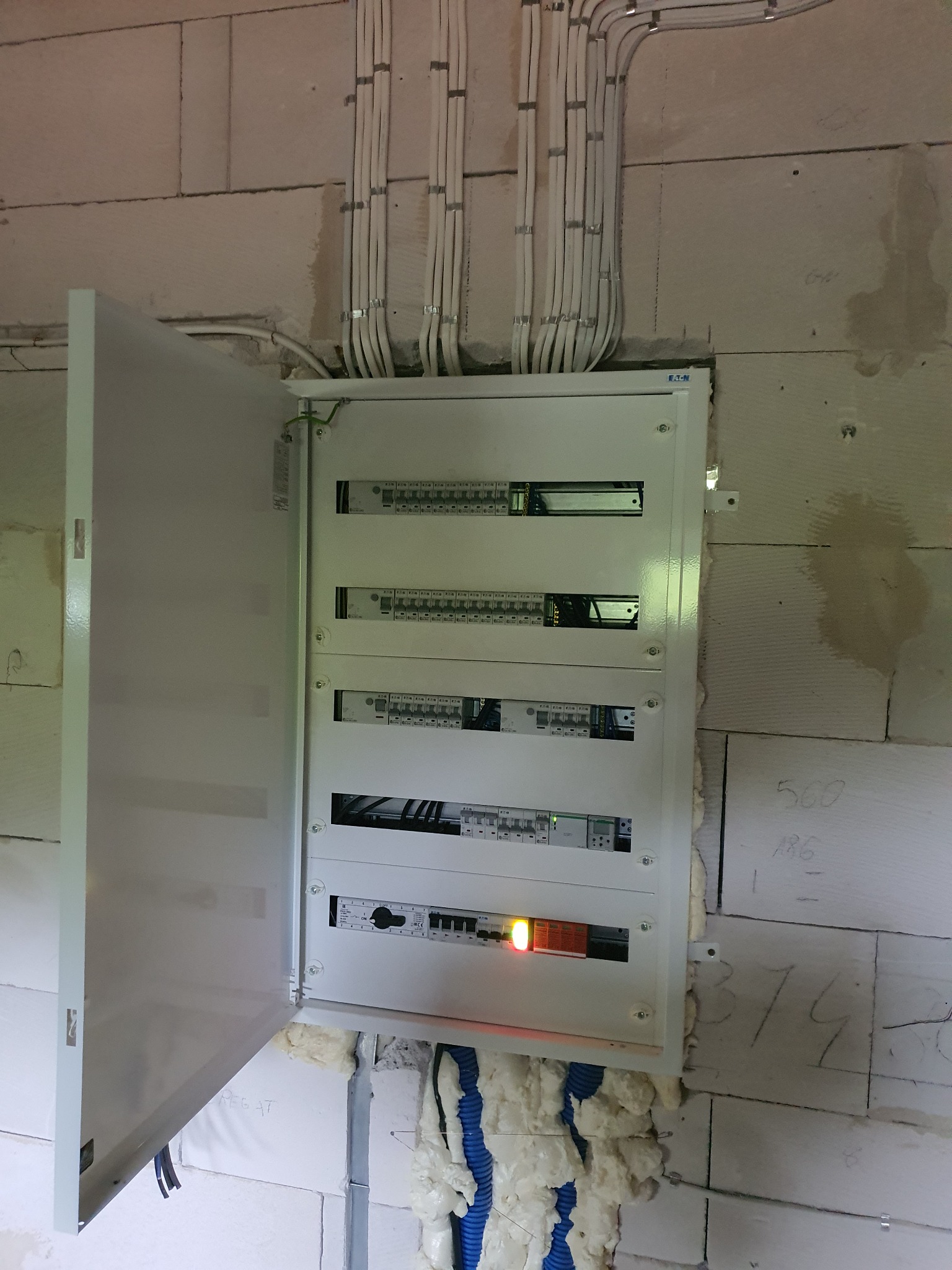 Otwarta skrzynka elektryczna z widocznymi bezpiecznikami i przewodami, zamontowana na surowej ścianie z betonu komórkowego, z widocznymi przewodami w peszlach prowadzonymi wzdłuż ściany.