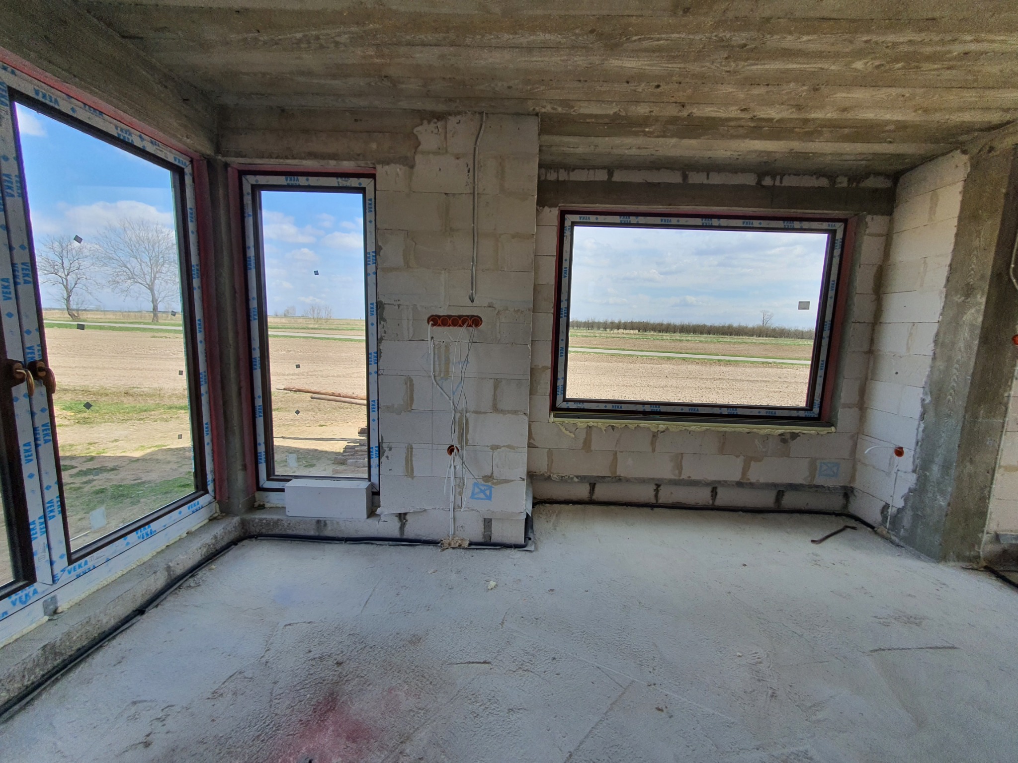 Wnętrze surowego domu w trakcie budowy z widocznymi przewodami elektrycznymi wystającymi ze ściany z pustaków, okna z widokiem na pole.