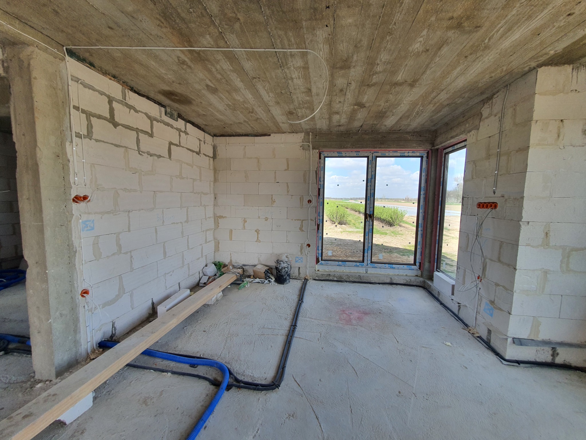 Wnętrze surowego domu w trakcie prac instalacyjnych; widoczne przewody elektryczne w ścianach z białego bloczka, rury instalacyjne na podłodze, okno z widokiem na zieleń.