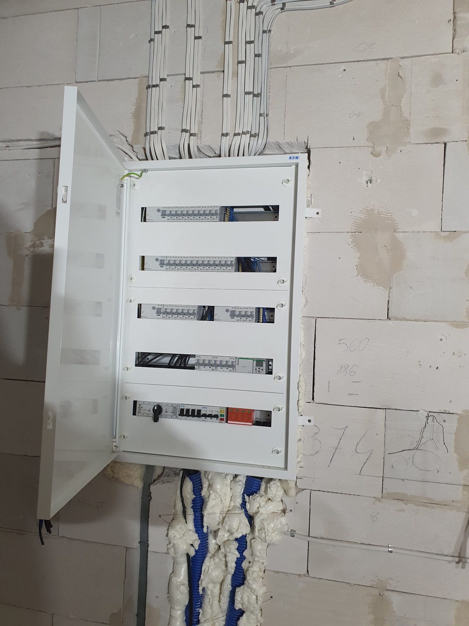 Otwarta skrzynka elektryczna z bezpiecznikami i wyłącznikami, zamontowana na surowej ścianie z betonu komórkowego z widocznymi przewodami w peszlach i pianką montażową.
