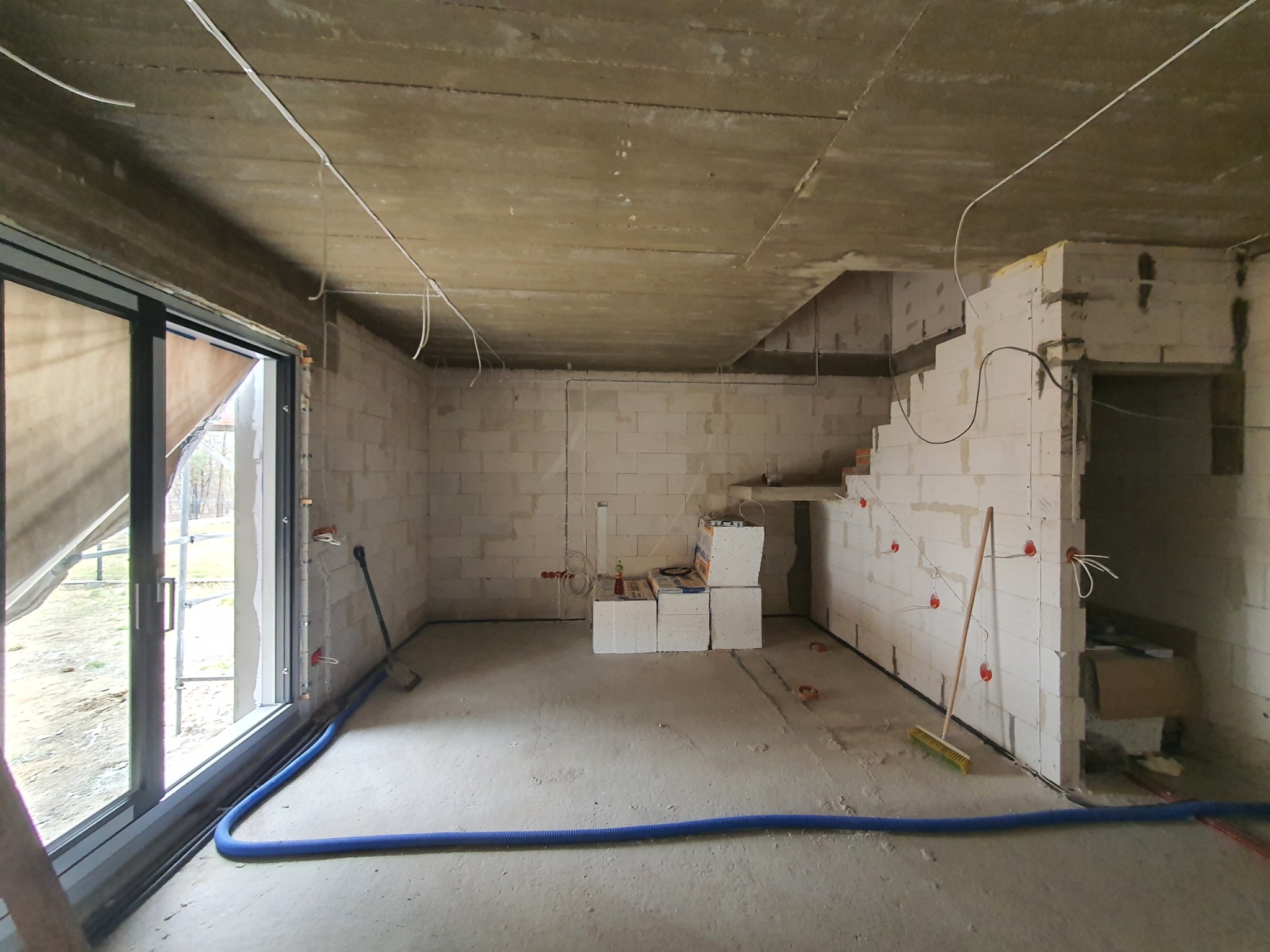 Surowy stan wnętrza z widocznymi przewodami elektrycznymi wystającymi ze ścian, nieotynkowane ściany z betonu komórkowego, niebieski peszel na podłodze, przygotowania do instalacji elektrycznej.