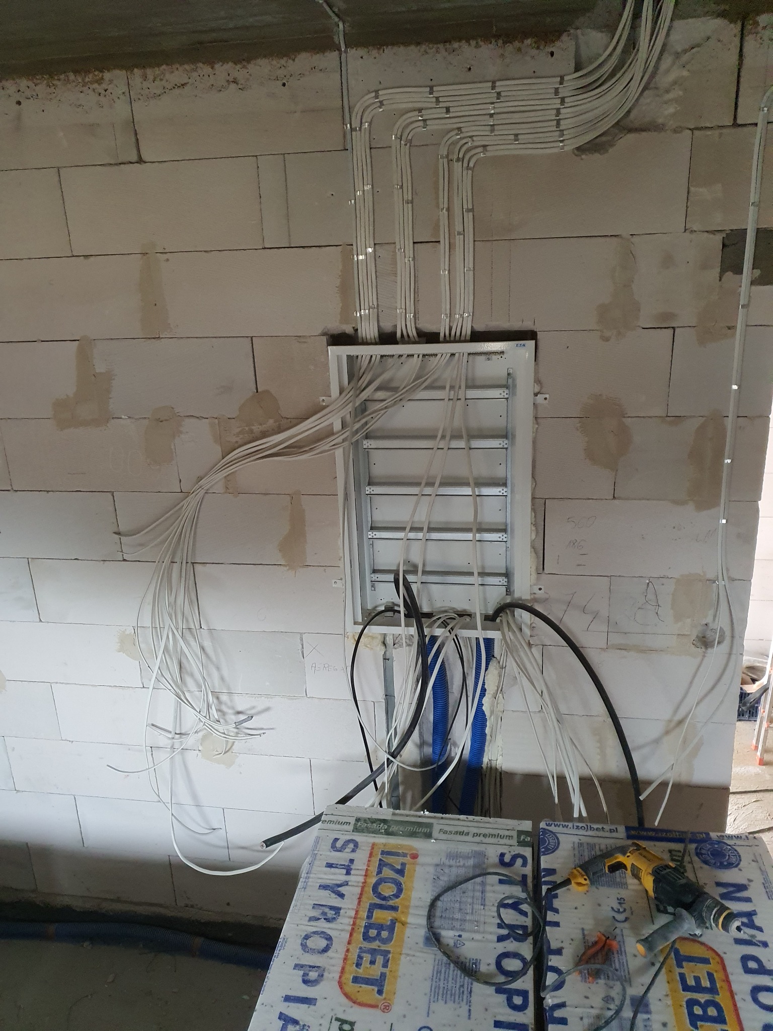 Rozdzielnica elektryczna z licznymi przewodami na surowej ścianie z betonu komórkowego, widoczne także narzędzia i materiały izolacyjne.