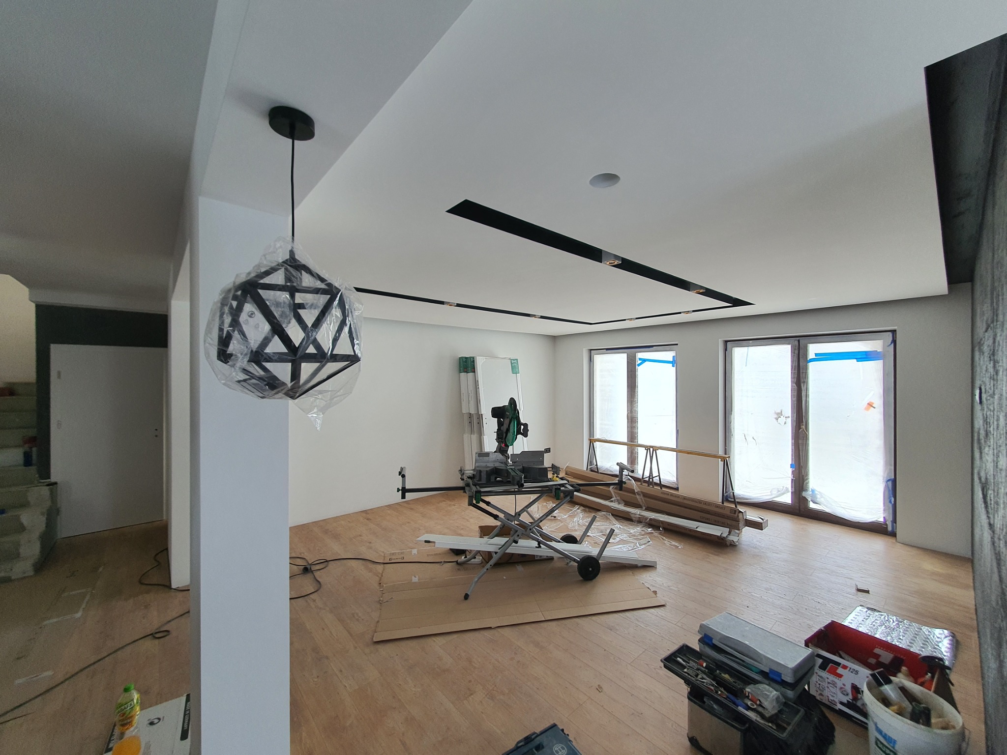 Wnętrze w trakcie wykańczania: podłoga z drewnianych paneli, narzędzia do cięcia listew przypodłogowych, okna zabezpieczone folią, wisząca lampa w geometrycznej obudowie, częściowo jeszcze w folii...
