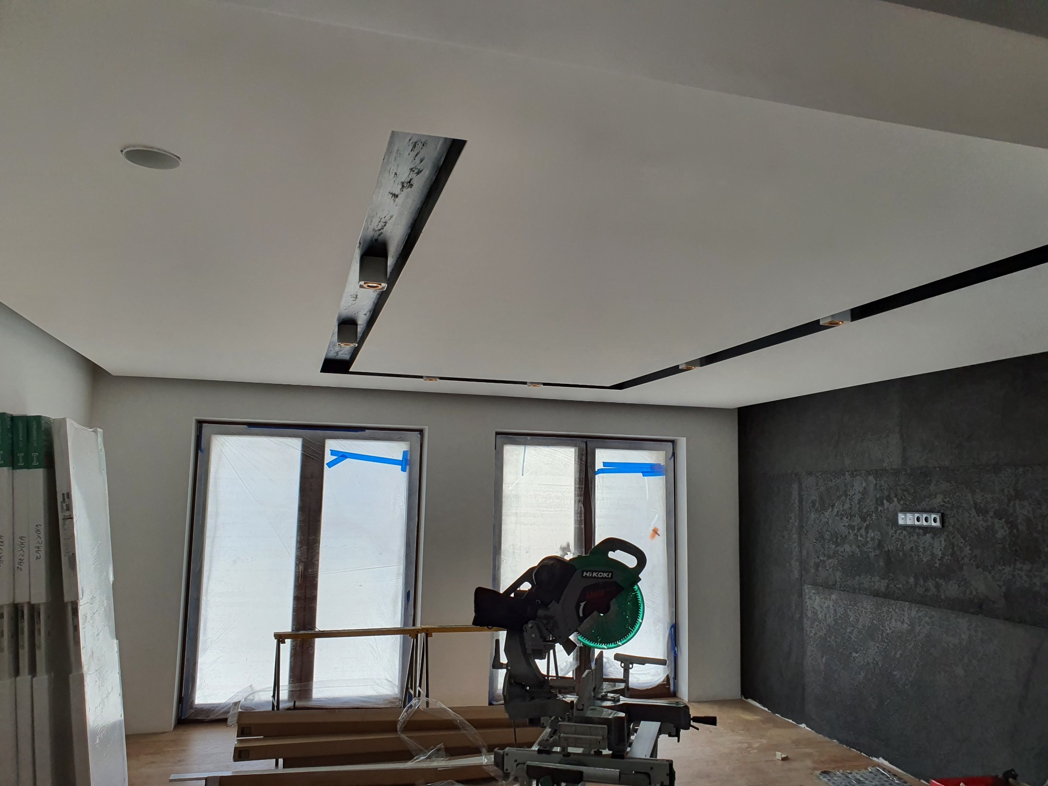 Wnętrze w trakcie remontu z widocznym oświetleniem sufitowym w czarnych listwach i wnękach, okna zabezpieczone folią, piła ukośnica na pierwszym planie.