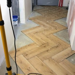 Andrzej Stachowski AMS - Układanie drewnopodobnych płytek podłogowych w jodełkę, widoczne krzyżyki dystansowe, narzędzia i lampa budowlana w pomieszczeniu w trakcie remontu.