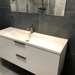 Nowoczesna łazienka: Biała, podwieszana szafka z umywalką i czarnymi uchwytami na tle szarych płytek imitujących kamień, widoczne minimalistyczne baterie i lustro.