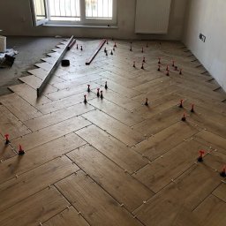 Andrzej Stachowski AMS - Układanie drewnopodobnych płytek podłogowych w jodełkę, widoczne poziomica, krzyżyki dystansowe i system poziomowania płytek z czerwonymi klinami.