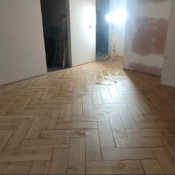 Andrzej Stachowski AMS - Ułożone panele podłogowe w jodełkę na mokro, widoczne gniazdka elektryczne na ścianie, pomieszczenie w trakcie prac wykończeniowych, lampa budowlana w tle.