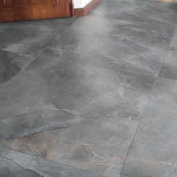 Andrzej Stachowski AMS - Szare płytki podłogowe imitujące kamień, ułożone w prostokątnym wzorze, widoczne w dolnej części drewniane drzwi i fragment kosza z wikliny.