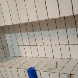 Andrzej Stachowski AMS - Świeżo ułożone, pionowe, kremowe płytki w łazience z wnęką, widoczne krzyżyki dystansowe, fragment niebieskiego narzędzia w rogu.