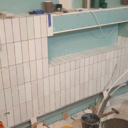 Andrzej Stachowski AMS - Ściana łazienki w trakcie układania białych, podłużnych płytek z widocznymi krzyżykami dystansowymi, z przygotowanym miejscem na półkę i instalacją hydrauliczną, narzędzia pracy w tle.