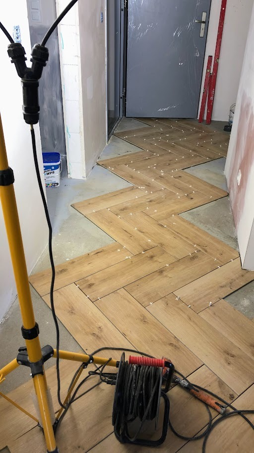Układanie drewnopodobnych płytek podłogowych w jodełkę, widoczne krzyżyki dystansowe, narzędzia i lampa budowlana w pomieszczeniu w trakcie remontu.