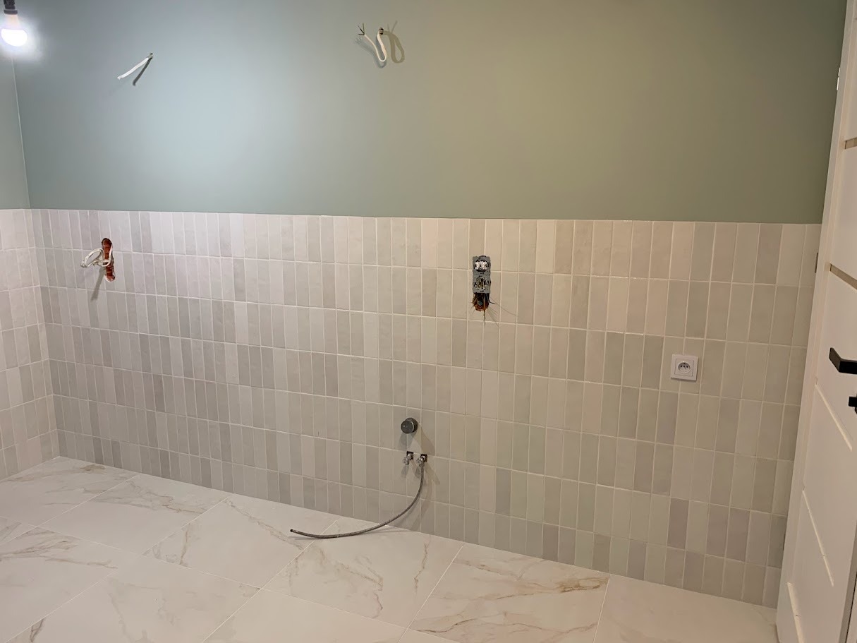 Łazienka w trakcie wykańczania: pionowe, beżowe płytki na ścianie do połowy wysokości, zielona farba powyżej, marmurowe płytki na podłodze, widoczne instalacje wodne i elektryczne przed montażem...