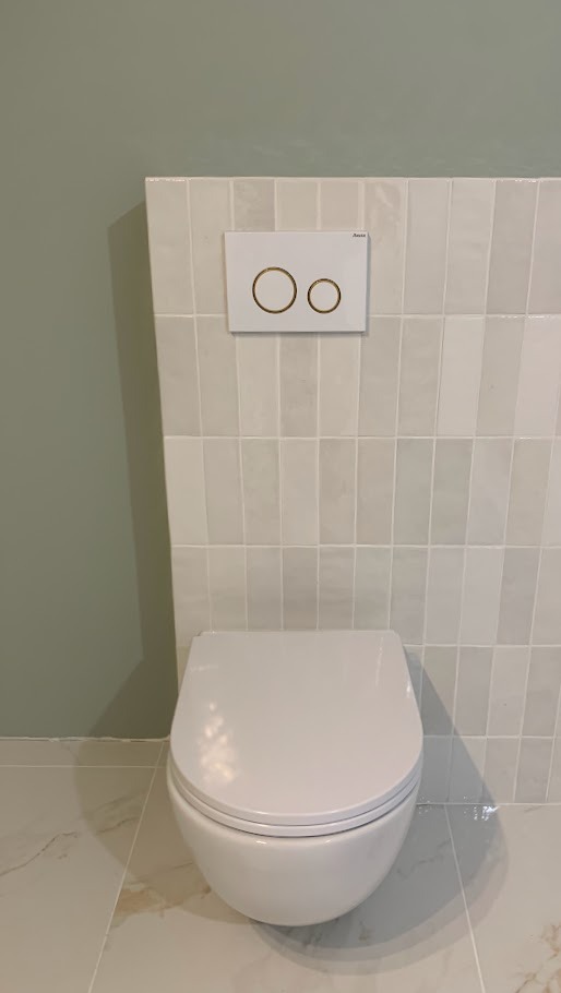 Podwieszana toaleta z białymi płytkami ułożonymi pionowo i przyciskiem spłukującym z dwoma złotymi okręgami, zamontowana na ścianie pomalowanej na oliwkowy kolor.