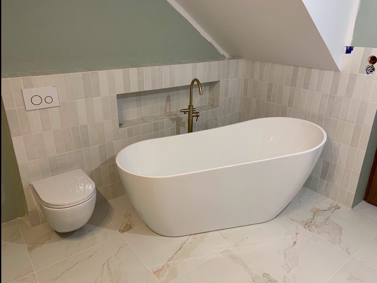 Nowoczesna łazienka w trakcie wykańczania: wolnostojąca biała wanna, złota bateria, podwieszana toaleta, płytki imitujące marmur na podłodze i pionowe, jasne płytki na ścianach, widoczne...