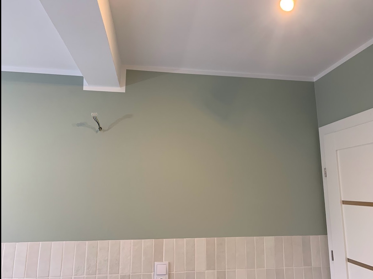 Ściana pomalowana na zielono-szary kolor z wystającymi przewodami elektrycznymi, biały sufit z listwą i fragment białych płytek na dole ściany oraz gniazdko elektryczne.