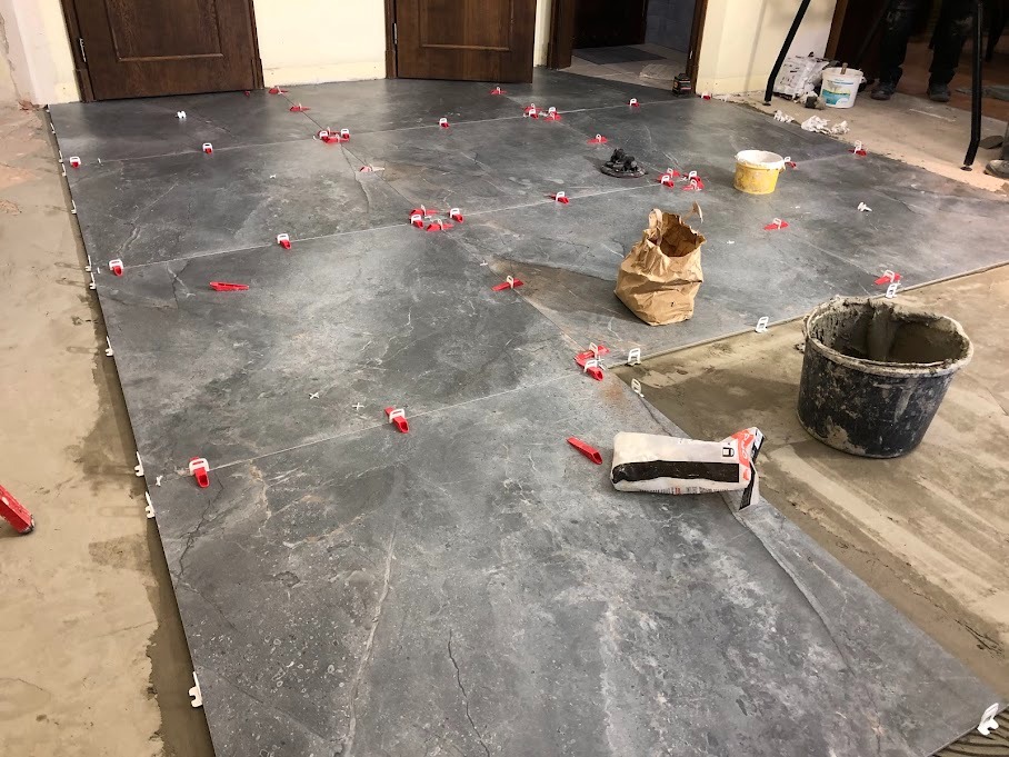 Układanie szarych płytek podłogowych z użyciem czerwonych klinów poziomujących, widoczny worek z klejem, wiadro z zaprawą i fragment niedokończonej podłogi.