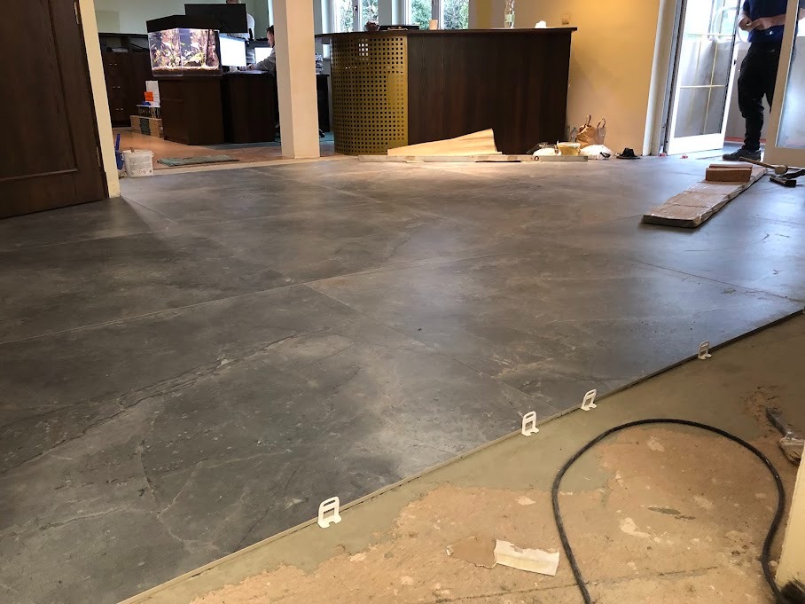 Układanie dużych, szarych płytek podłogowych w przestrzeni biurowej, widoczne białe klipsy poziomujące i fragment nieukończonej podłogi, w tle akwarium i recepcja.