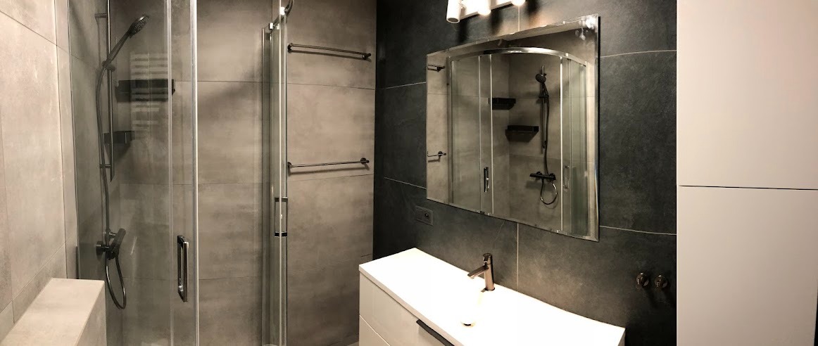 Nowoczesna łazienka z kabiną prysznicową z czarną armaturą, dużymi szarymi płytkami, białą umywalką nablatową i lustrem.