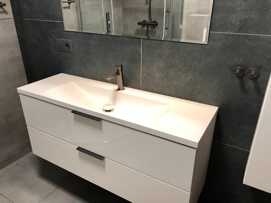 Nowoczesna łazienka: Biała, podwieszana szafka z umywalką i czarnymi uchwytami na tle szarych płytek imitujących kamień, widoczne minimalistyczne baterie i lustro.