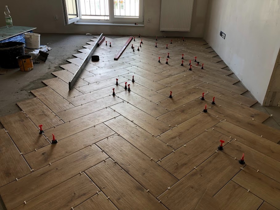 Układanie drewnopodobnych płytek podłogowych w jodełkę, widoczne poziomica, krzyżyki dystansowe i system poziomowania płytek z czerwonymi klinami.