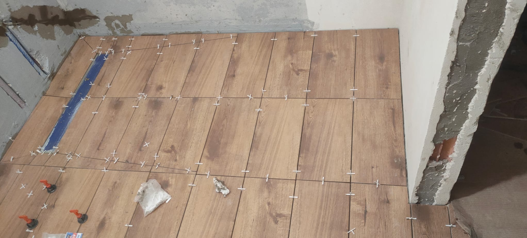 Układanie płytek imitujących drewno na podłodze w łazience, widoczne krzyżyki dystansowe, poziomica wzdłuż ściany i niezakończone wykończenie ściany.
