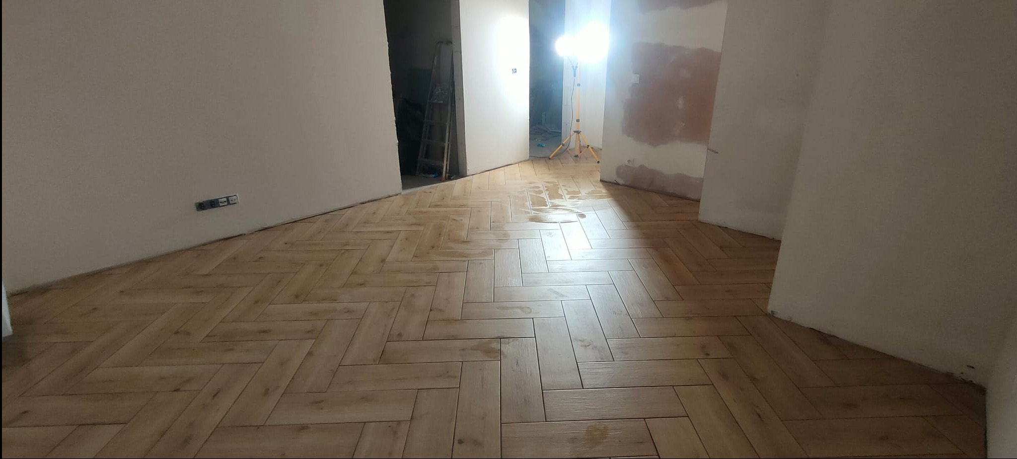 Ułożone panele podłogowe w jodełkę na mokro, widoczne gniazdka elektryczne na ścianie, pomieszczenie w trakcie prac wykończeniowych, lampa budowlana w tle.
