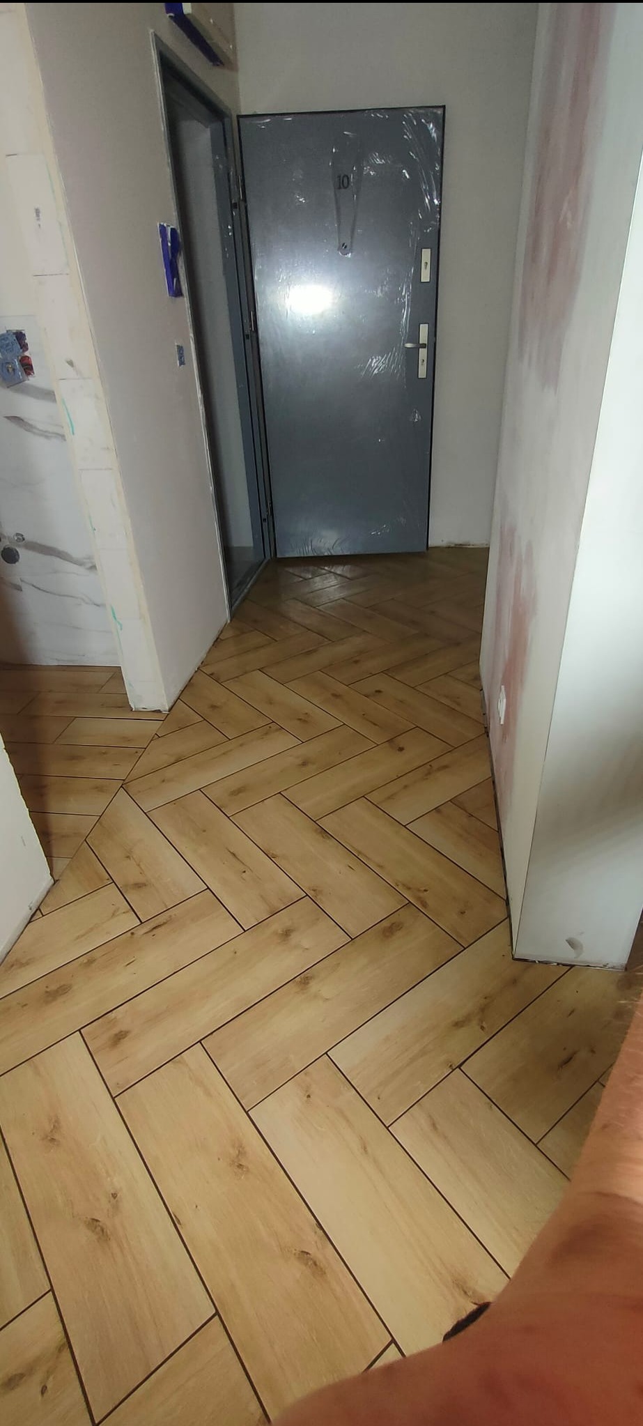 Nowo ułożona podłoga z paneli imitujących drewno w jodełkę, widoczna szara, zabezpieczona folią ochronną, metalowa futryna drzwi wejściowych do mieszkania z numerem 10, ściany w trakcie...