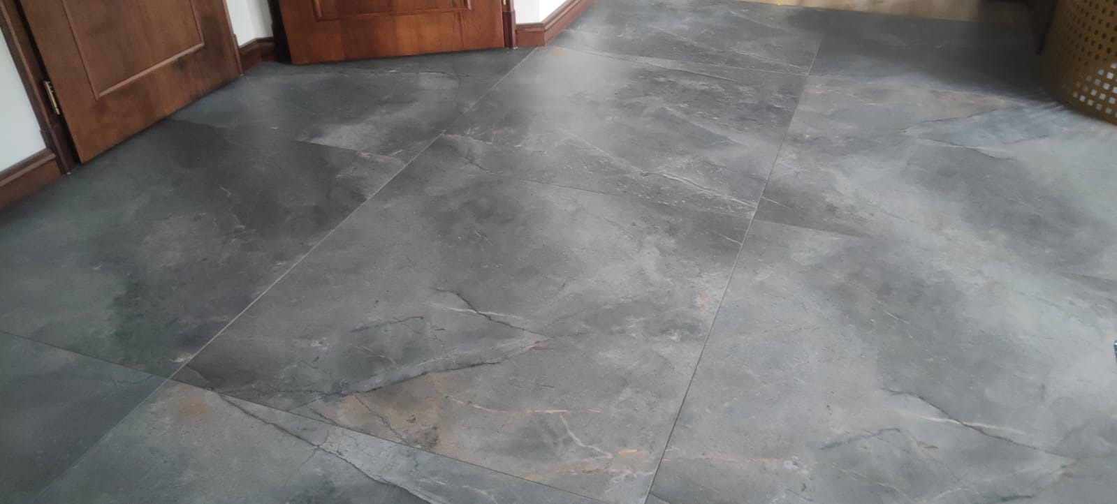 Szare płytki podłogowe imitujące kamień, ułożone w prostokątnym wzorze, widoczne w dolnej części drewniane drzwi i fragment kosza z wikliny.