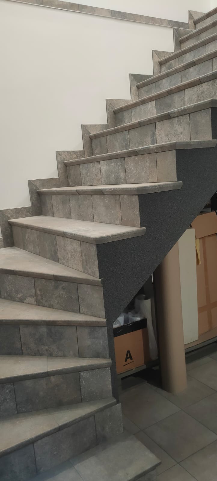 Wykończone schody wewnętrzne z szarych płytek imitujących kamień, widoczne z dołu, z szarą, chropowatą ścianą pod spodem, częściowo zasłonięte kartonami i rolką papieru.