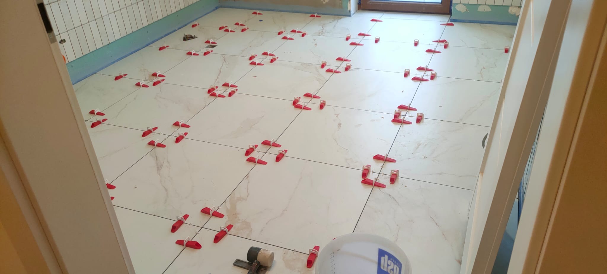 Białe płytki podłogowe z marmurowym wzorem, układane z użyciem czerwonych klipsów poziomujących, widok z perspektywy wejścia do łazienki, w tle fragment ściany z białymi płytkami.