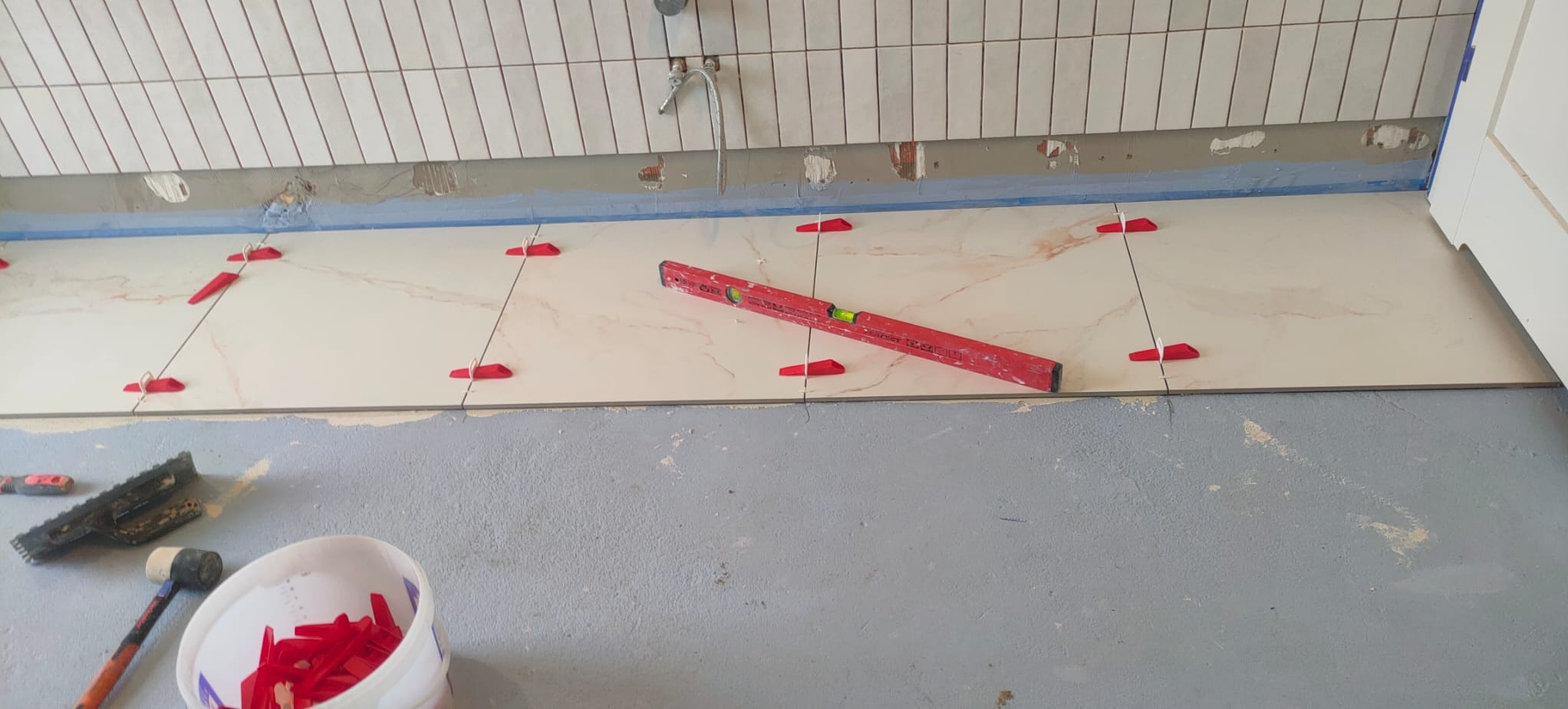 Układanie płytek podłogowych z użyciem poziomicy i czerwonych klinów dystansowych; widoczne narzędzia: kielnia, gumowy młotek i wiadro z klinami.