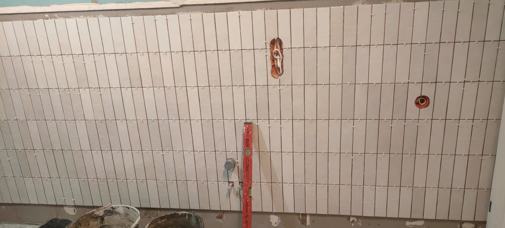 Ściana łazienki w trakcie układania podłużnych, białych płytek z krzyżykami dystansowymi, widoczne otwory na instalacje i poziomica, dwa wiadra na podłodze.