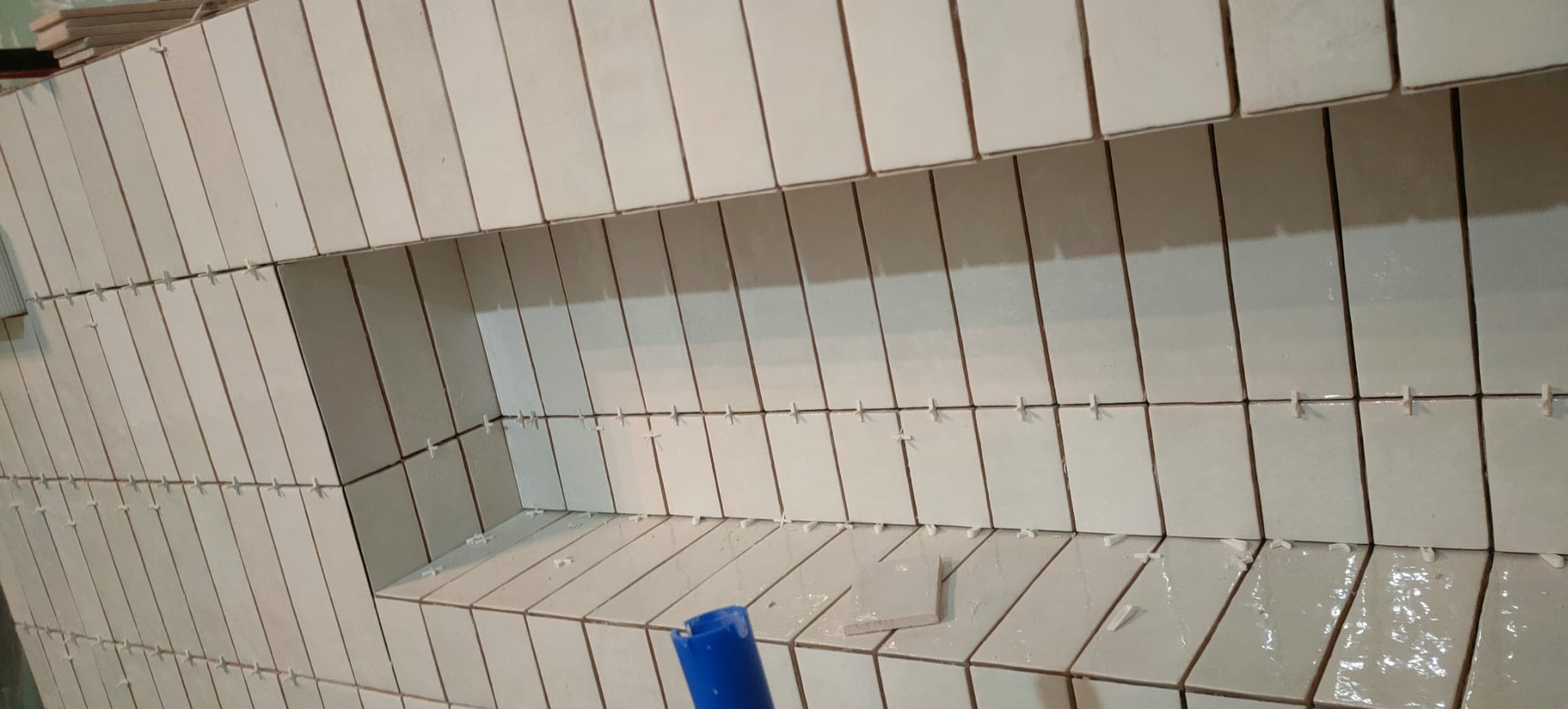 Świeżo ułożone, pionowe, kremowe płytki w łazience z wnęką, widoczne krzyżyki dystansowe, fragment niebieskiego narzędzia w rogu.