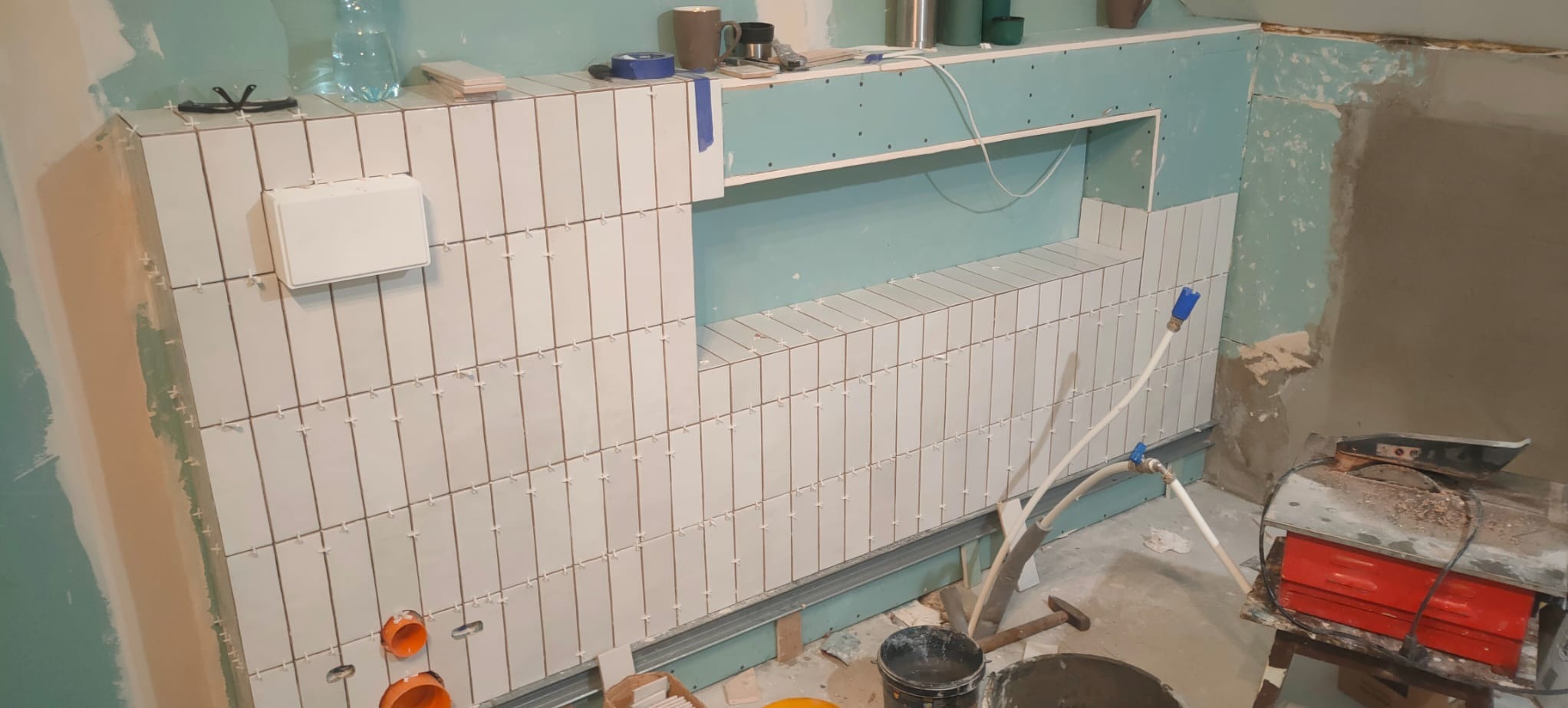 Ściana łazienki w trakcie układania białych, podłużnych płytek z widocznymi krzyżykami dystansowymi, z przygotowanym miejscem na półkę i instalacją hydrauliczną, narzędzia pracy w tle.