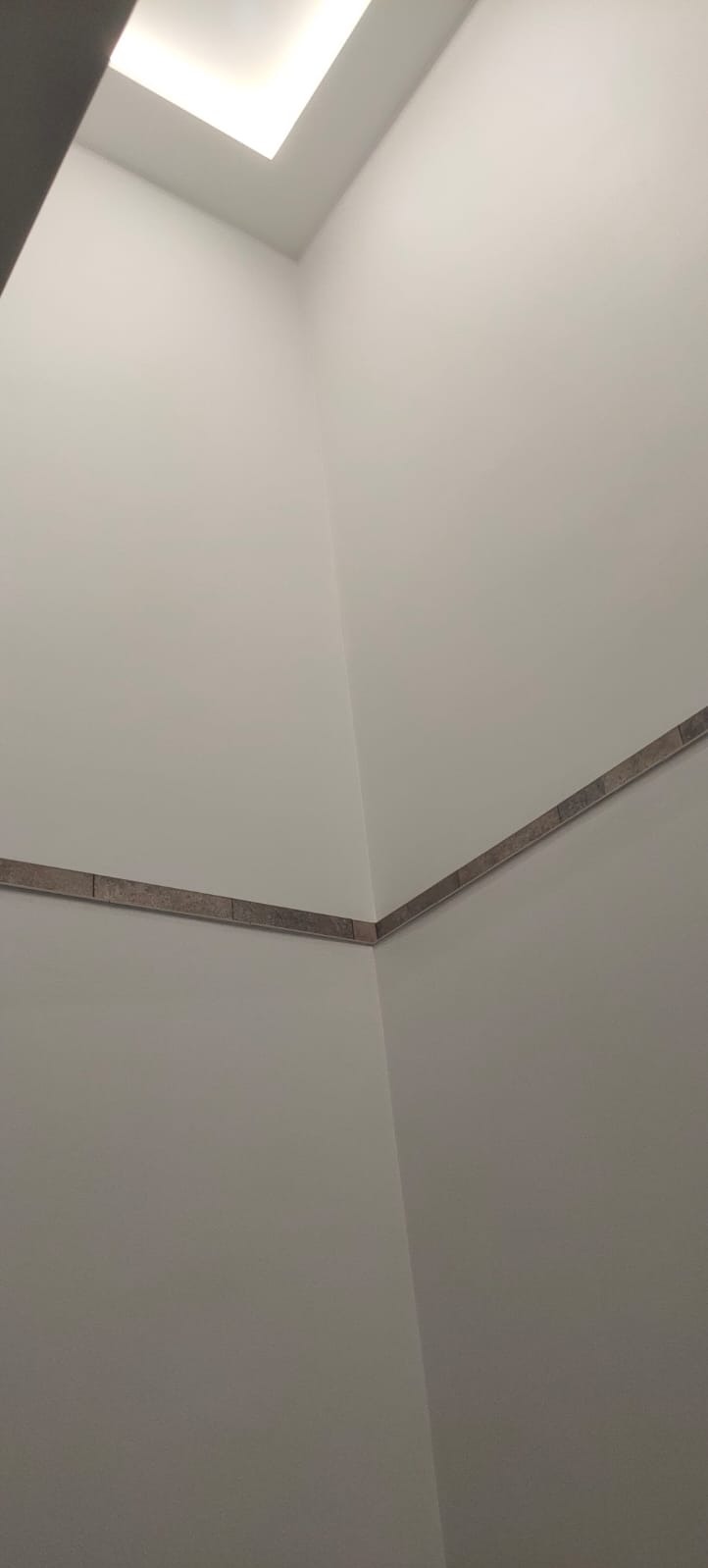 Fragment ściany w jasnym pomieszczeniu z wykończeniem w postaci wąskiego pasa płytek dekoracyjnych pod sufitem i oświetleniem sufitowym.