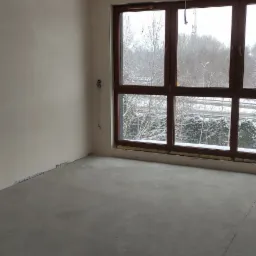 Surowe wnętrze pokoju po nałożeniu tynków, widoczne ślady po pracach wykończeniowych na podłodze, duże okno z widokiem na zaśnieżone drzewa.