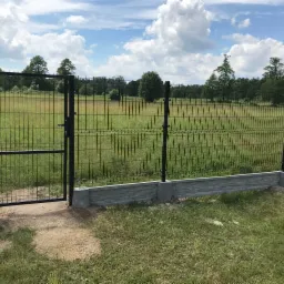 Ciemna, metalowa siatka ogrodzeniowa z furtką na betonowej podmurówce, widok na łąkę i las w tle pod błękitnym niebem z chmurami.