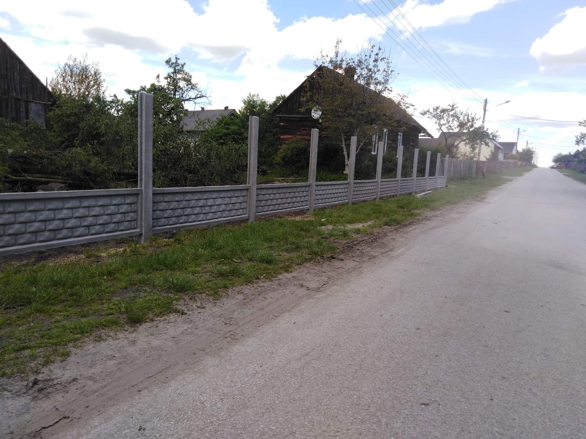 Szary, betonowy płot panelowy z prefabrykatów, z widocznymi słupkami, oddzielający posesję od wiejskiej drogi asfaltowej, w tle zabudowania gospodarcze i drzewa.