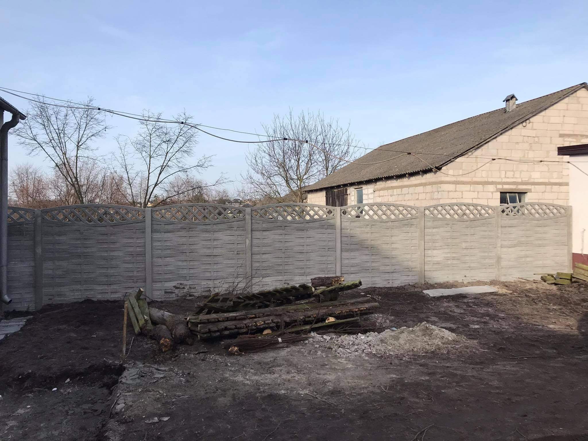 Nowo postawione, betonowe ogrodzenie panelowe z dekoracyjnym wzorem w górnej części, widoczne na tle starego, murowanego budynku gospodarczego z kominem, teren wokół ogrodzenia jeszcze...